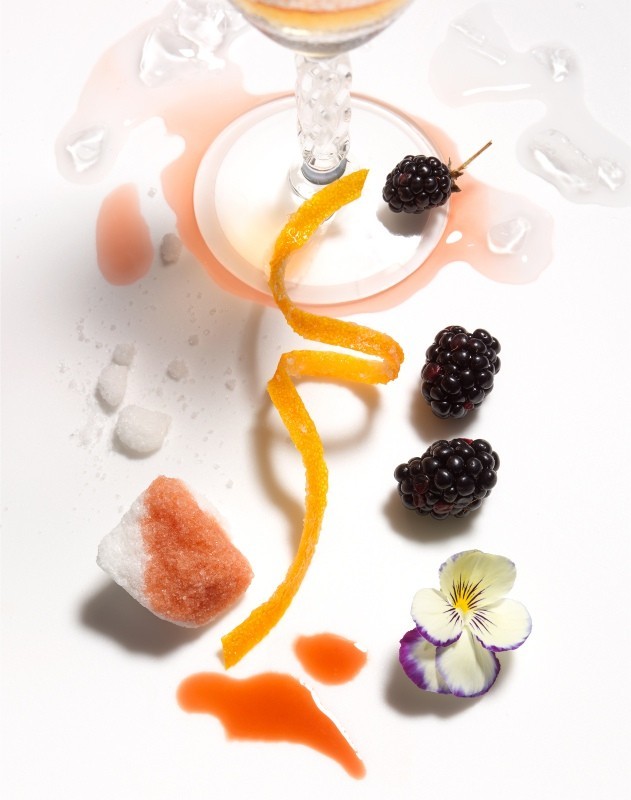 Cocktail glass with an orange peel twist, blackberries, sugar cubes, and edible flower on a white background, top-down view.