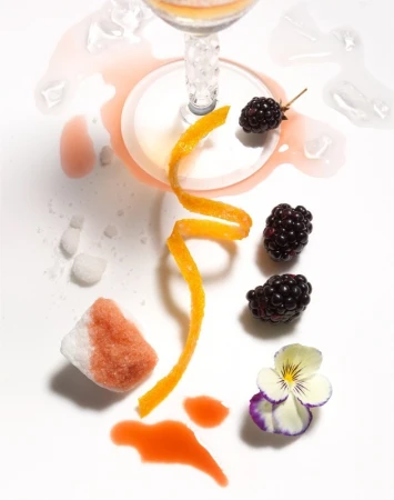 Cocktail glass with an orange peel twist, blackberries, sugar cubes, and edible flower on a white background, top-down view.