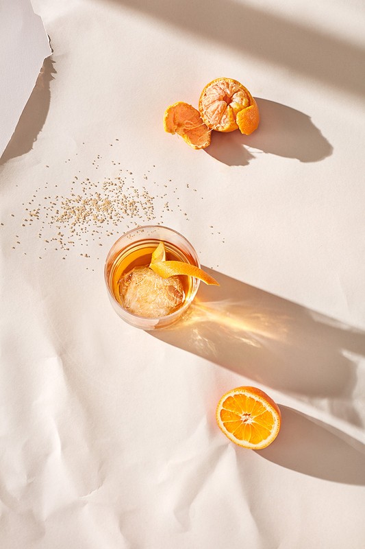 Overhead shot of an orange cocktail garnished with a peel, an orange half, a peeled orange, and scattered sugar on white paper.