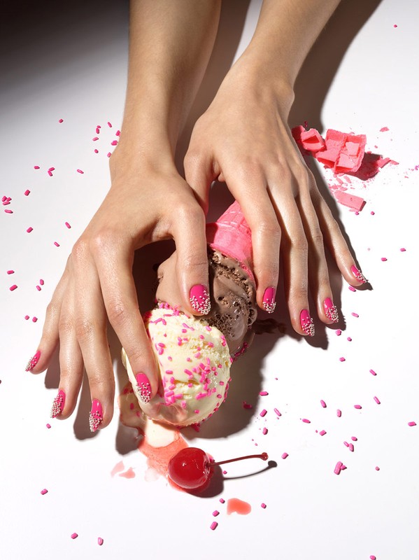 Close-up of two hands with pink manicured nails holding an ice cream cone with three scoops of ice cream, topped with pink sprinkles.
