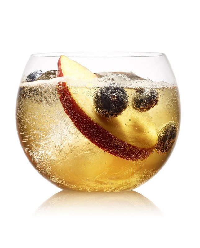 Close-up of a clear glass filled with a golden drink, ice cubes, blueberries, and an apple slice, against a bright white background.