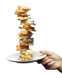 A hand holding a white plate balancing a towering stack of cheese and crackers against a white background.