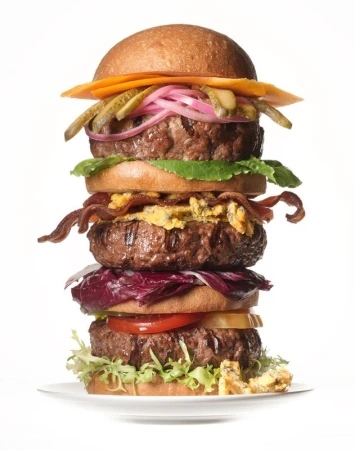 A towering multi-patty cheeseburger with bacon, pickles, and lettuce on a white plate against a plain background.