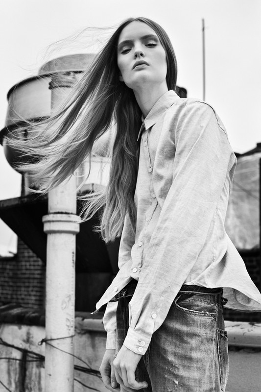 Monochrome portrait of a woman with long hair, wearing a collared shirt and jeans, posing on a rooftop with industrial elements.