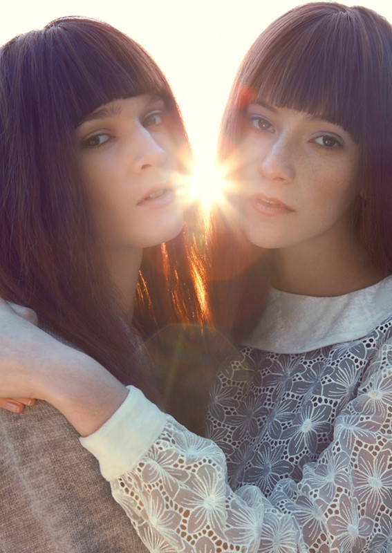 Close-up of two young adult women with dark hair and bangs embracing in a brightly lit outdoor setting. Focus on their faces.