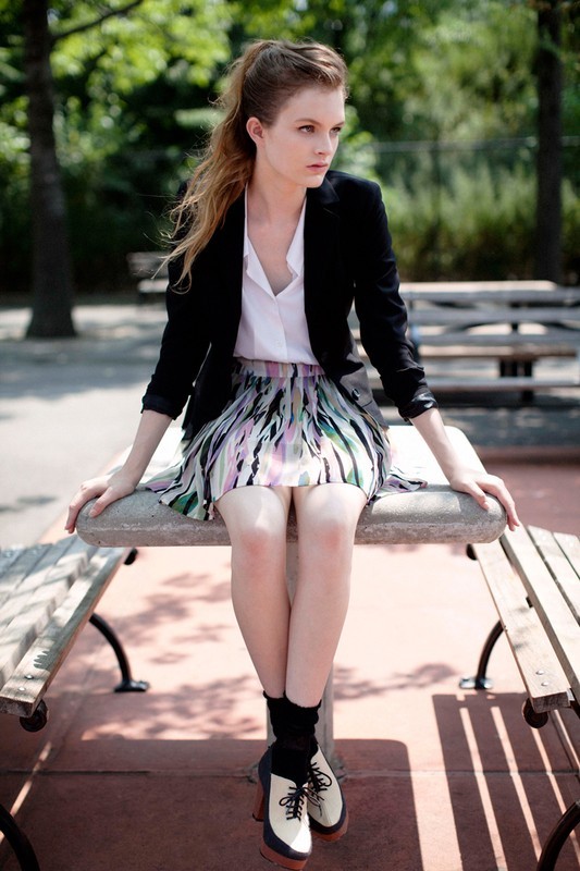 Young woman in fashionable outfit sits on park bench with trees in the background, wearing a jacket, skirt, socks, and platform shoes.