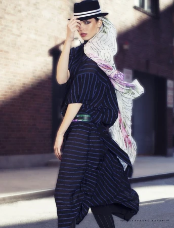Model in dark fedora and striped outfit with flowing floral scarf standing outdoors on a city street.
