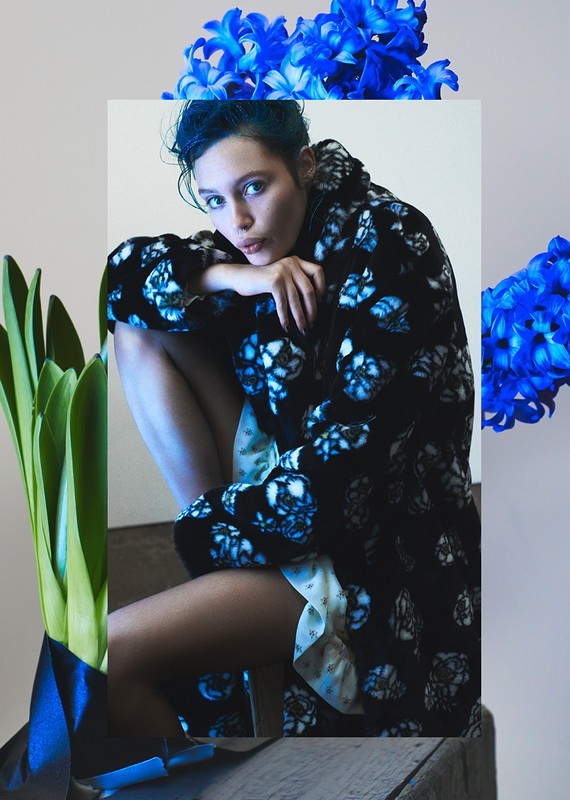 Portrait of a young woman in a patterned robe sitting indoors next to blue hyacinth flowers and green foliage with a neutral background.