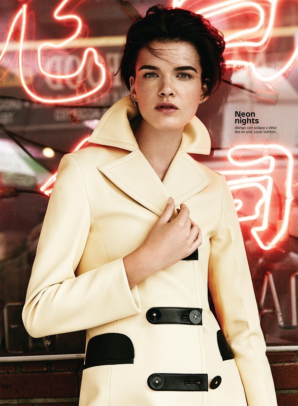 A fair-skinned woman with short hair poses in a pale-yellow coat, standing in front of neon lights in a fashion-style photograph.