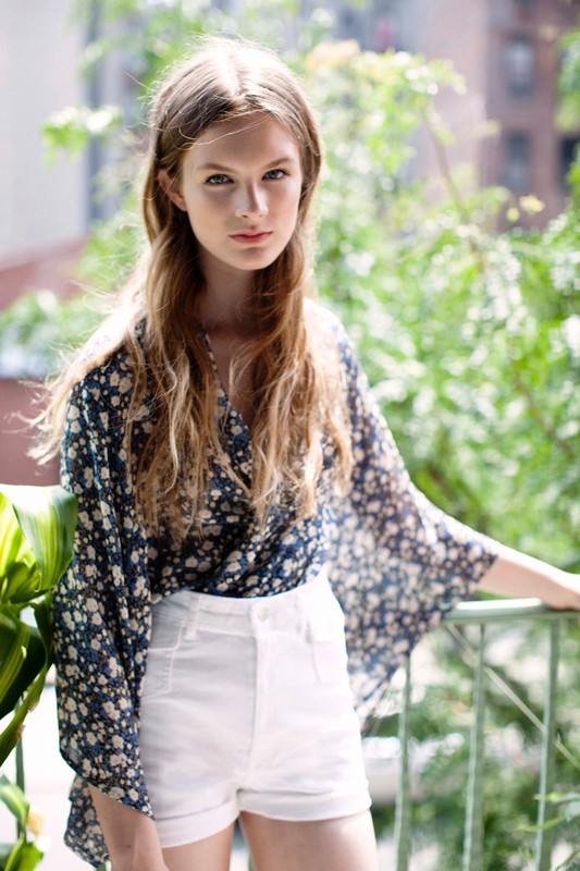 Blonde woman in a floral top and white shorts leans against a railing on a balcony with green foliage in the background.