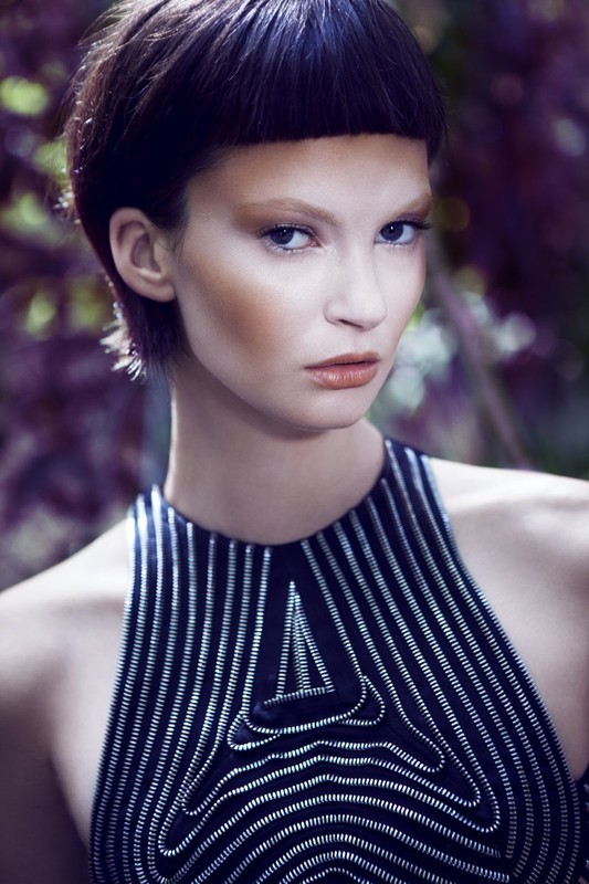 Portrait of a woman with a short dark haircut and a futuristic-patterned sleeveless top, posing against a blurred natural background with greenery.
