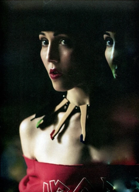 Portrait of a young woman with a hat and a necklace made of legs, the background is dark, and there is another partial face.