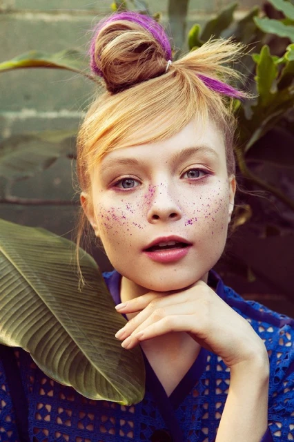 Close-up of a young woman with blonde hair styled in a bun with purple streaks, adorned with glitter on her face, holding a large leaf.