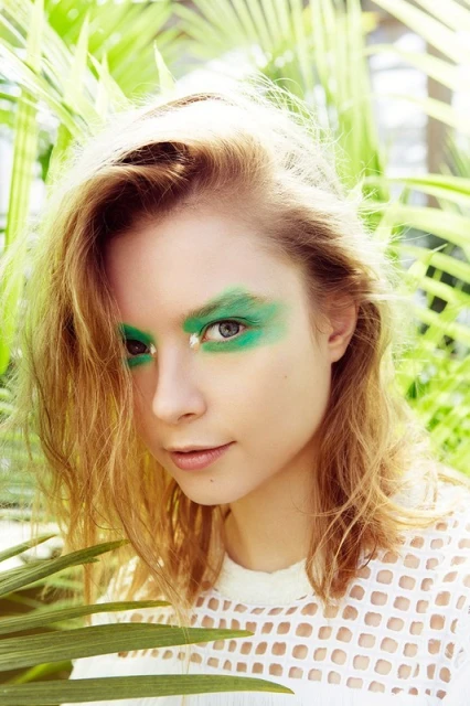 Close-up of a young woman with blonde hair and green eyeshadow posing outdoors among green plants, wearing a white mesh top.