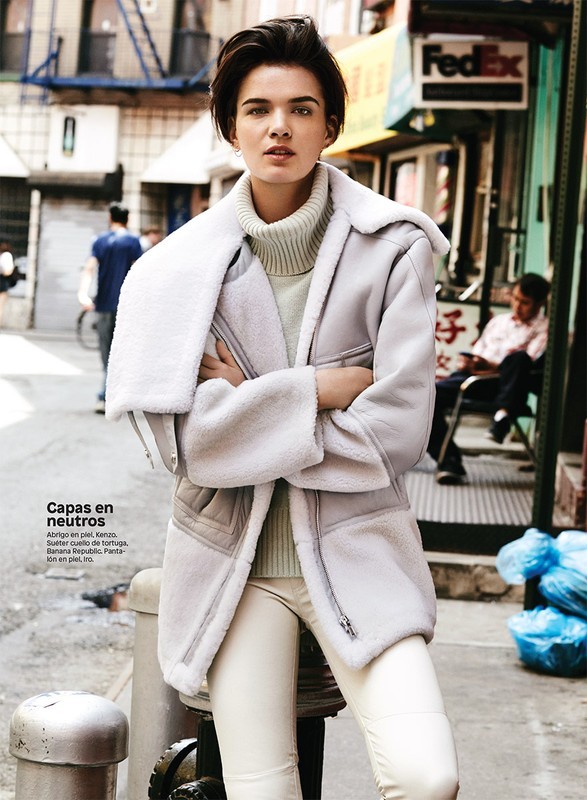 Woman in stylish grey shearling coat, turtleneck, and white pants stands on urban street with FedEx sign in the background.