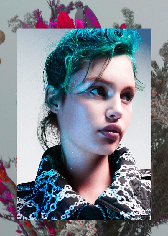 Close-up of a woman with blue-green hair wearing a chain-patterned jacket against a blurred floral background in a fashion shot.
