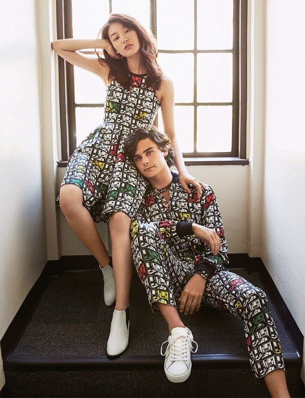 A stylish couple in matching patterned outfits poses indoors, with a woman seated and a man reclining on stairs near a window.