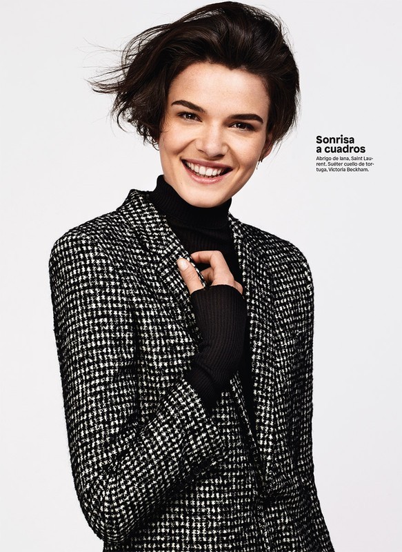 Smiling woman in a black and white patterned blazer and black turtleneck sweater poses against a plain light gray background.