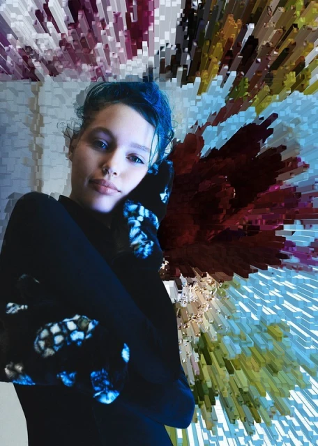 Portrait of a woman with blue hair and dark clothing, posing in front of a digitally altered background with geometric shapes.