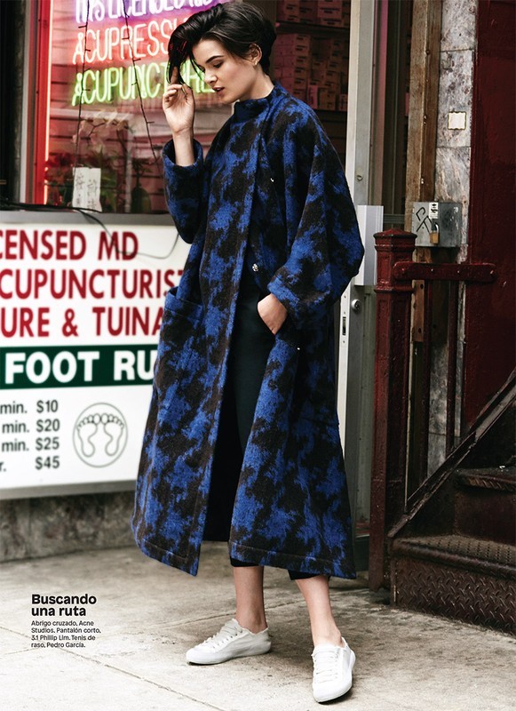 Woman in a long blue coat with black pattern, black pants, and white sneakers standing on an urban sidewalk in front of a storefront.