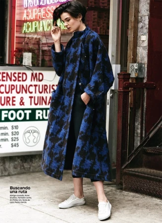 Woman in a long blue coat with black pattern, black pants, and white sneakers standing on an urban sidewalk in front of a storefront.