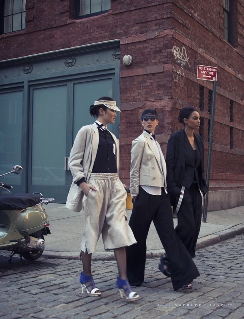 Three women pose outdoors on a city street in blazers, button-down shirts, and trousers, next to a scooter, urban fashion editorial.