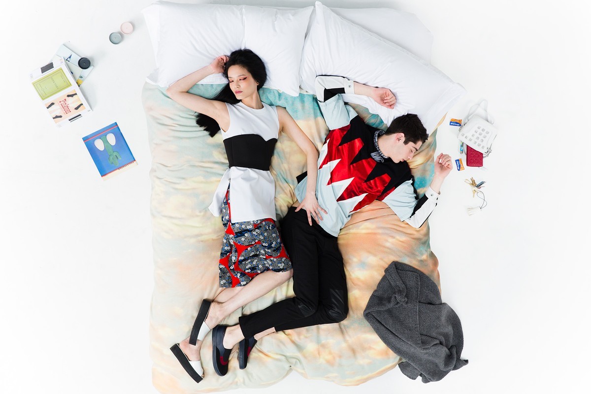 An overhead view of a man and woman lying on a colourful patterned bed with white pillows against a plain white background.