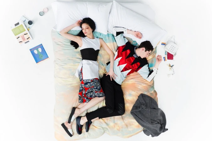 An overhead view of a man and woman lying on a colourful patterned bed with white pillows against a plain white background.