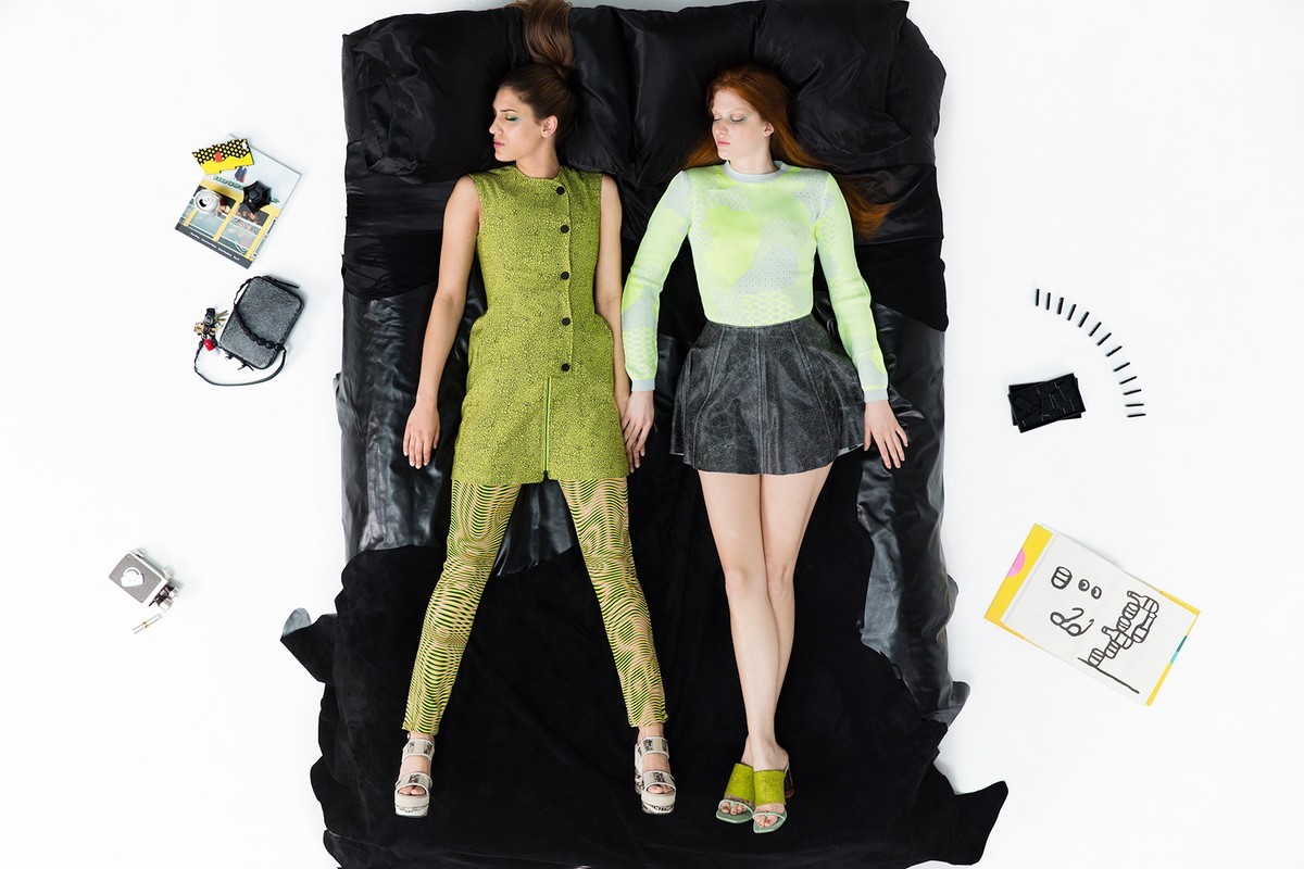 Two female models lying flat on a black bedding set surrounded by scattered accessories against a white background.