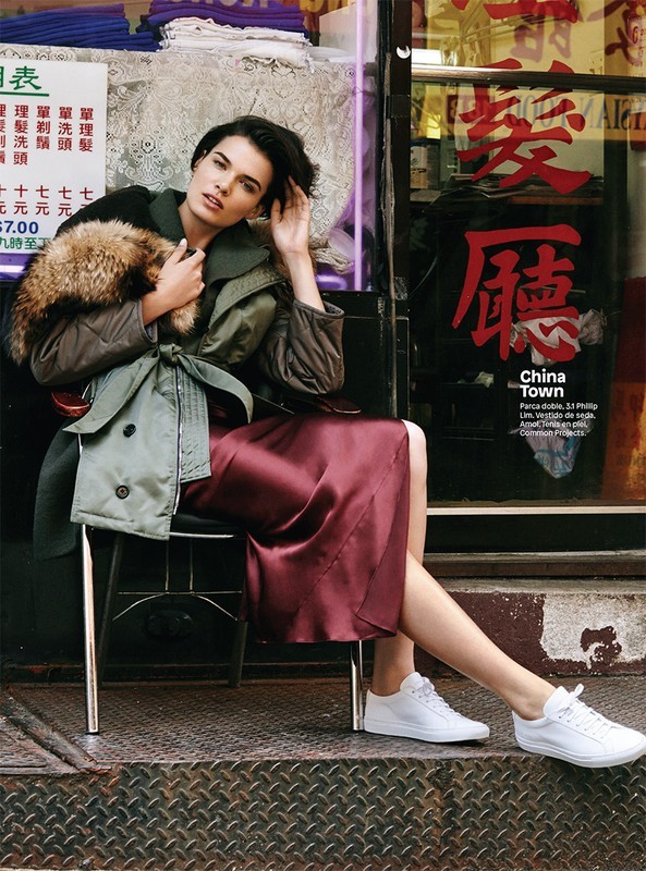 Elegant woman in stylish outerwear and a maroon dress sits in front of a shop with Chinese lettering. She's wearing white sneakers.