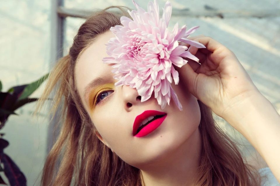 Close-up portrait of a woman with red lipstick and yellow eyeshadow holding a pink flower covering part of her face, indoors.
