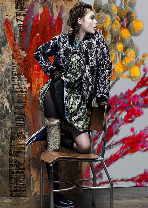 A stylish young woman poses on a chair in a patterned jacket and dress against a vibrant abstract background. Fashion editorial shot.