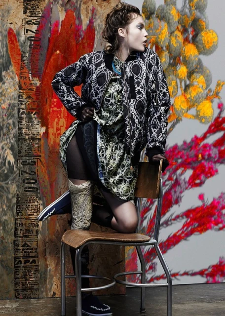 A stylish young woman poses on a chair in a patterned jacket and dress against a vibrant abstract background. Fashion editorial shot.