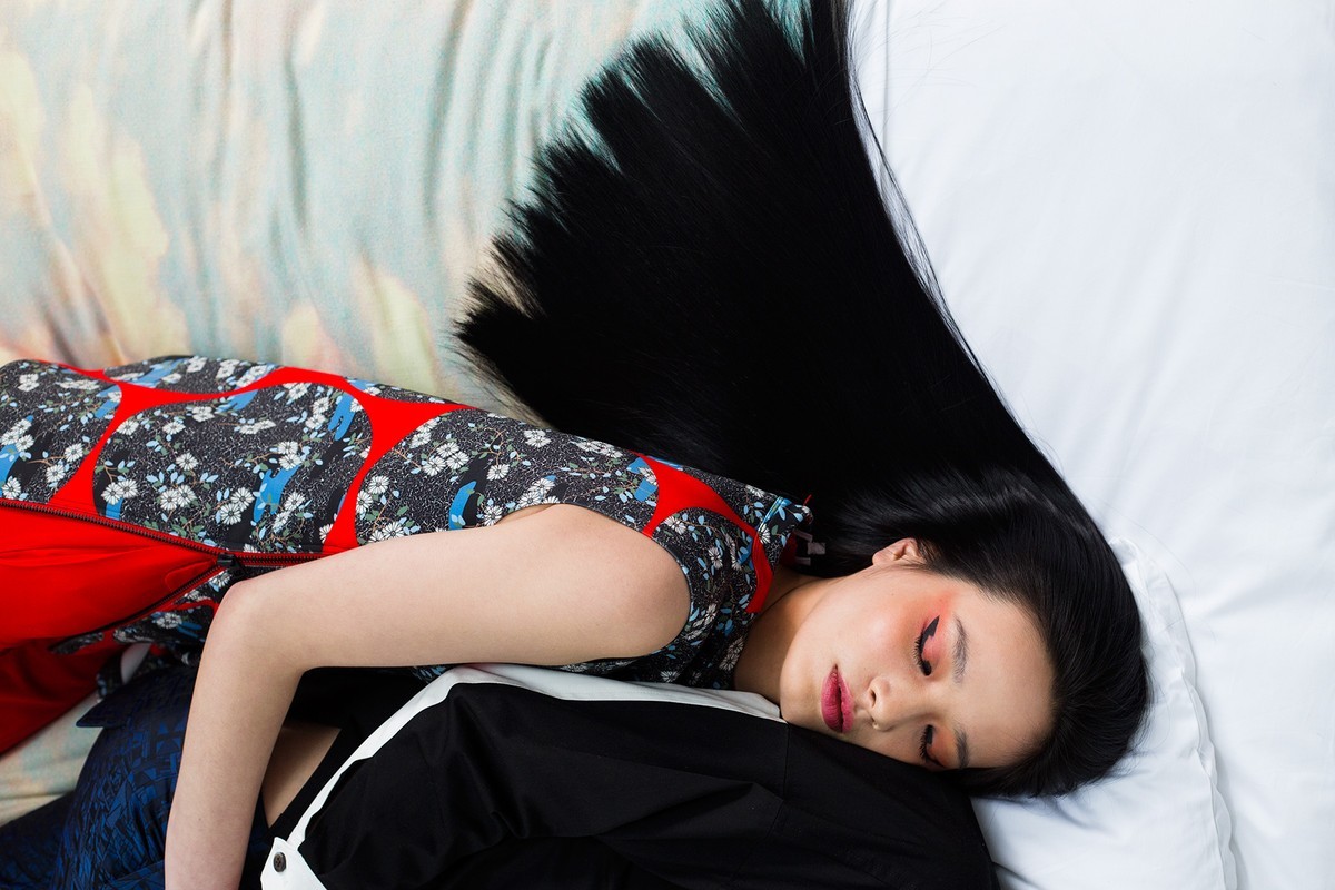 Woman with long black hair rests her face on a white pillow, wearing a red and black floral top and red pants, against a patterned backdrop.