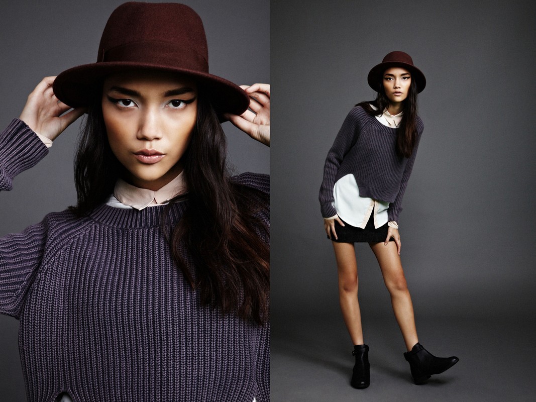 A model wearing a dark brown fedora, purple knit sweater, white collared shirt, and black skirt against a grey studio backdrop.