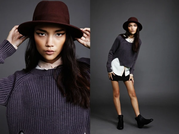 A model wearing a dark brown fedora, purple knit sweater, white collared shirt, and black skirt against a grey studio backdrop.