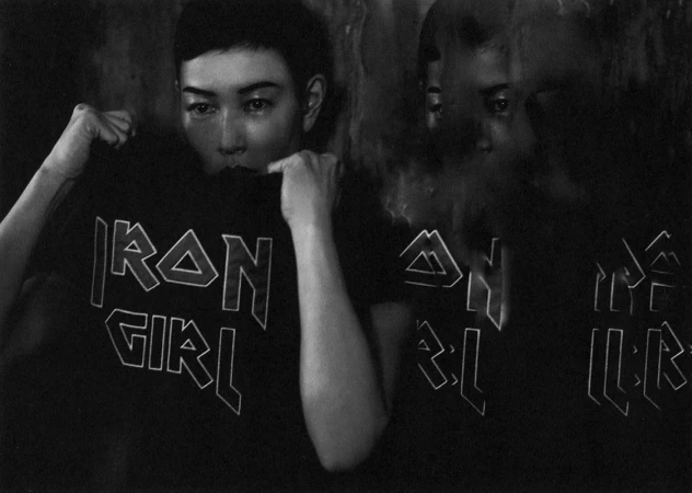 Monochrome multiple exposure portrait of a woman pulling up a black t-shirt with stylized lettering against a dark textured wall.
