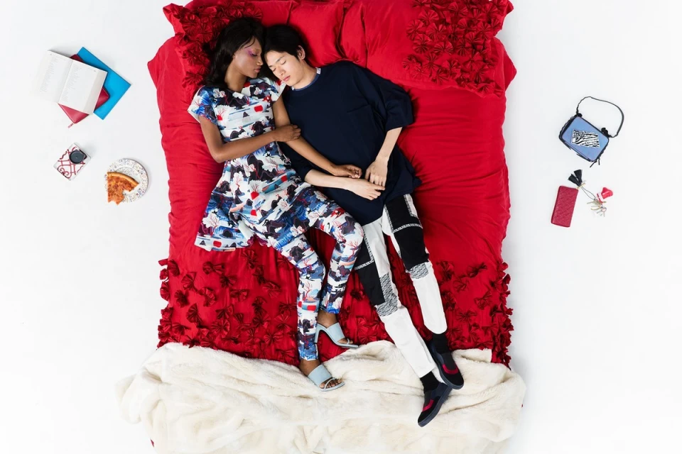 Two people lie embraced on a vibrant red bed with books and accessories scattered around, creating a cozy and fashionable indoor scene.
