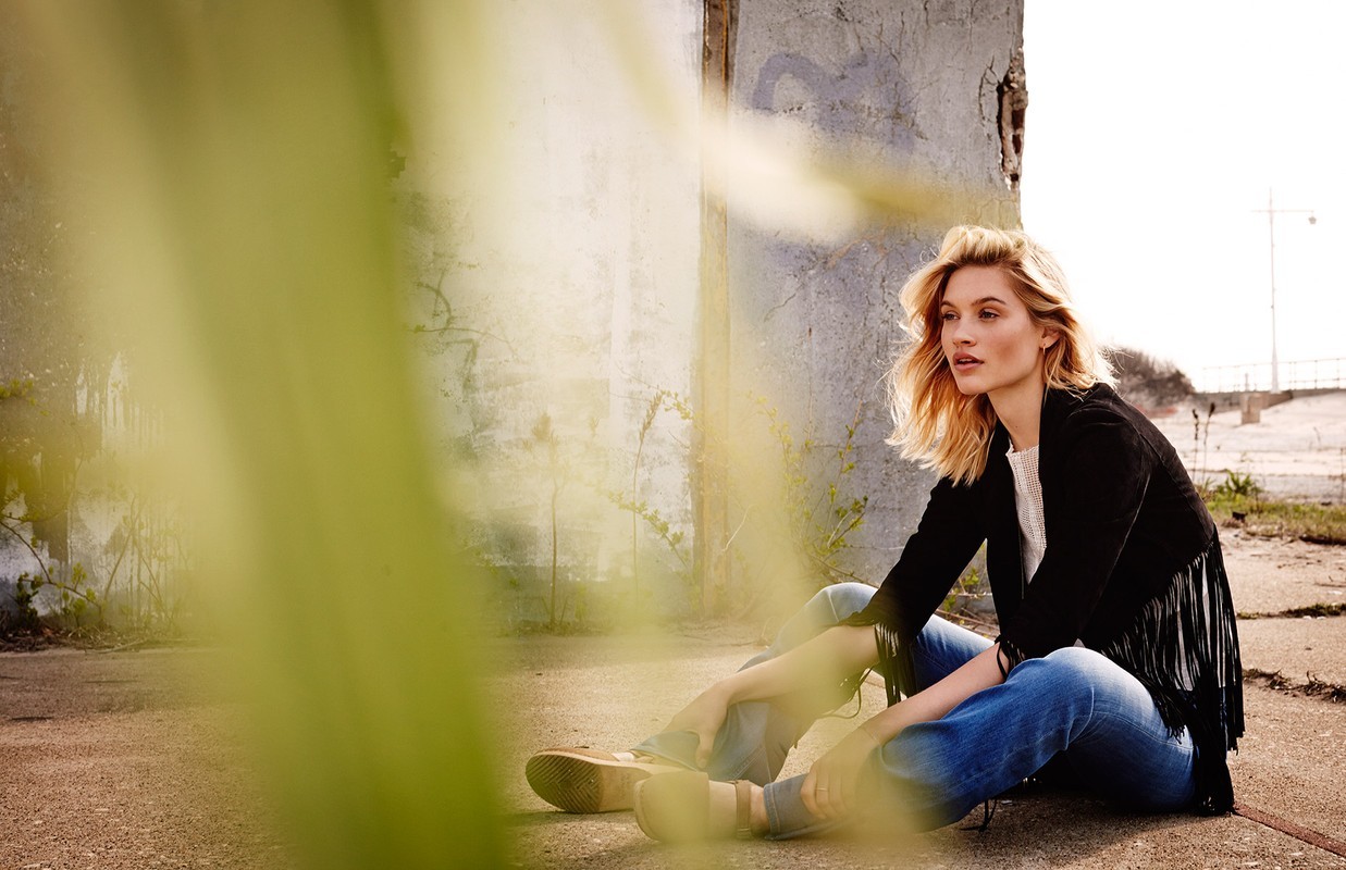 A blond woman sits on the ground in blue jeans and a black fringed jacket, posing against a weathered wall with greenery in the foreground.
