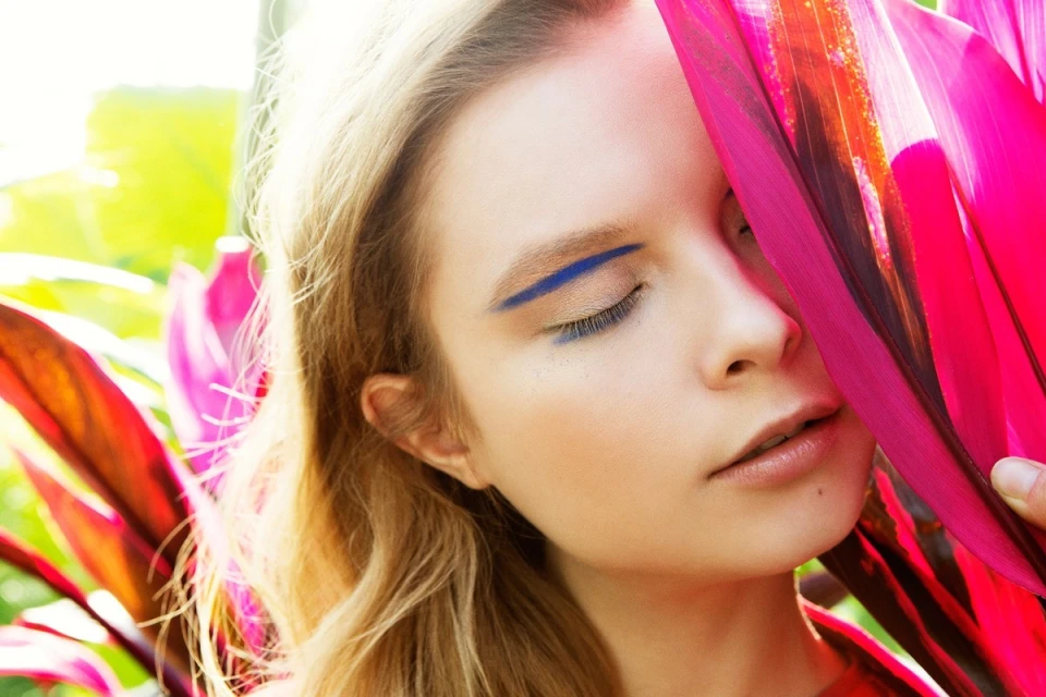 Close-up portrait of a young adult woman with blue eye makeup, surrounded by vibrant magenta tropical leaves in an outdoor setting.