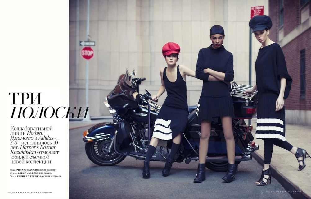Three fashion models in black outfits pose next to a motorcycle on a city street, showcasing a blend of urban style and edgy fashion.