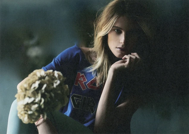 Close-up portrait of a woman with blonde hair wearing a blue graphic t-shirt, holding a bouquet of dried flowers.