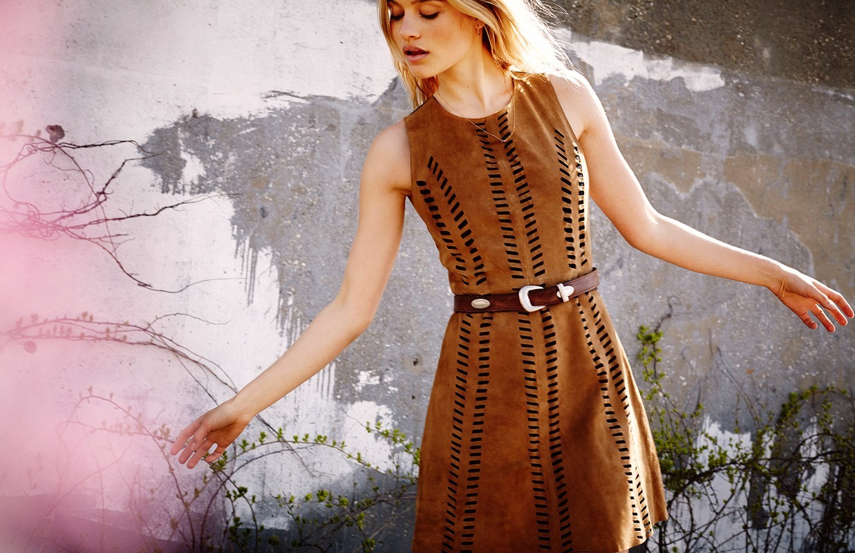 A woman poses in a sleeveless brown dress with a belt outdoors against a concrete wall with vines, highlighted by soft natural light.