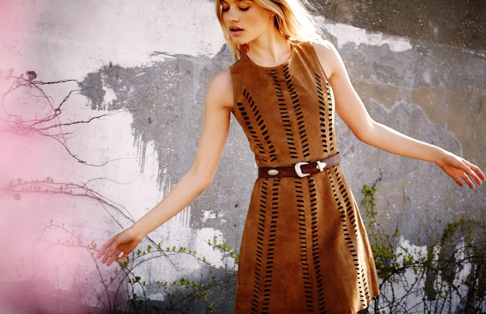 A woman poses in a sleeveless brown dress with a belt outdoors against a concrete wall with vines, highlighted by soft natural light.