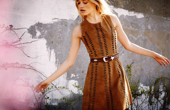 A woman poses in a sleeveless brown dress with a belt outdoors against a concrete wall with vines, highlighted by soft natural light.
