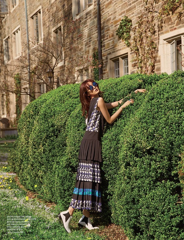 A stylish woman poses near greenery by a stone building, wearing a sleeveless top, pleated skirt, ankle boots, and sunglasses, outdoors.