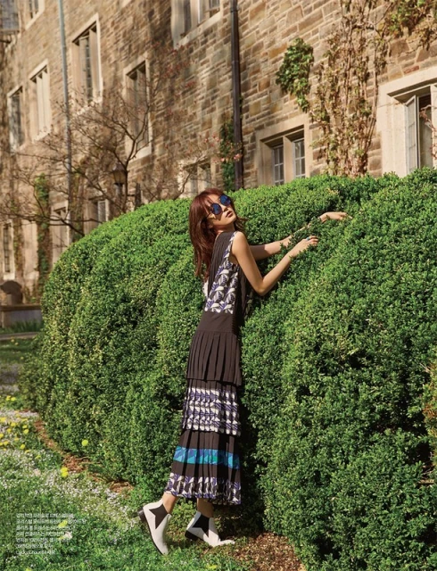 A stylish woman poses near greenery by a stone building, wearing a sleeveless top, pleated skirt, ankle boots, and sunglasses, outdoors.