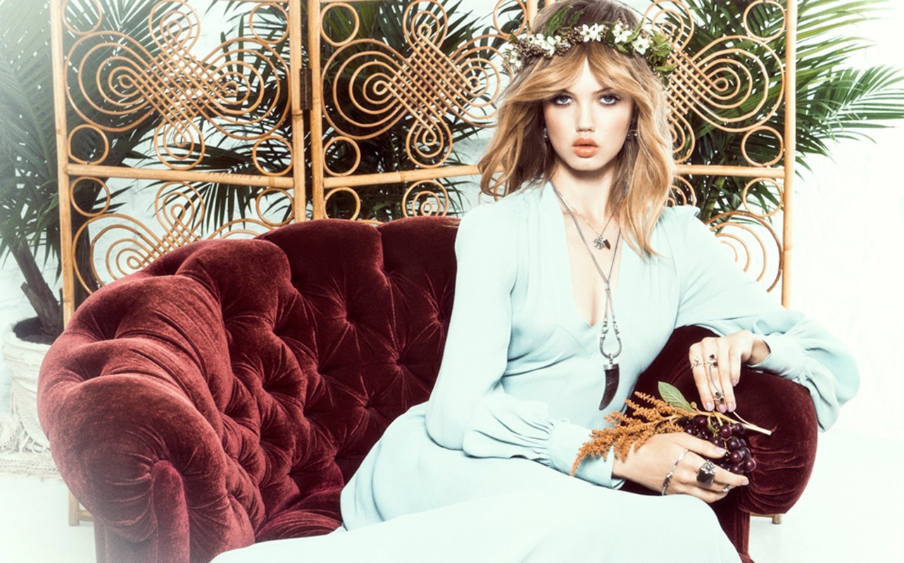 Portrait of a blonde woman with a floral crown sitting on a velvet sofa in front of a bamboo screen with a fern plant.