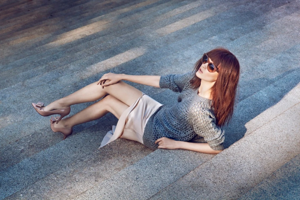 Elegant woman in sunglasses and grey sweater poses on concrete stairs, wearing a light pink skirt and matching high-heeled shoes.