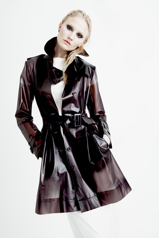 Fashionable woman in a glossy brown trench coat posing in a bright white studio setting, showcasing modern style and clothing design.